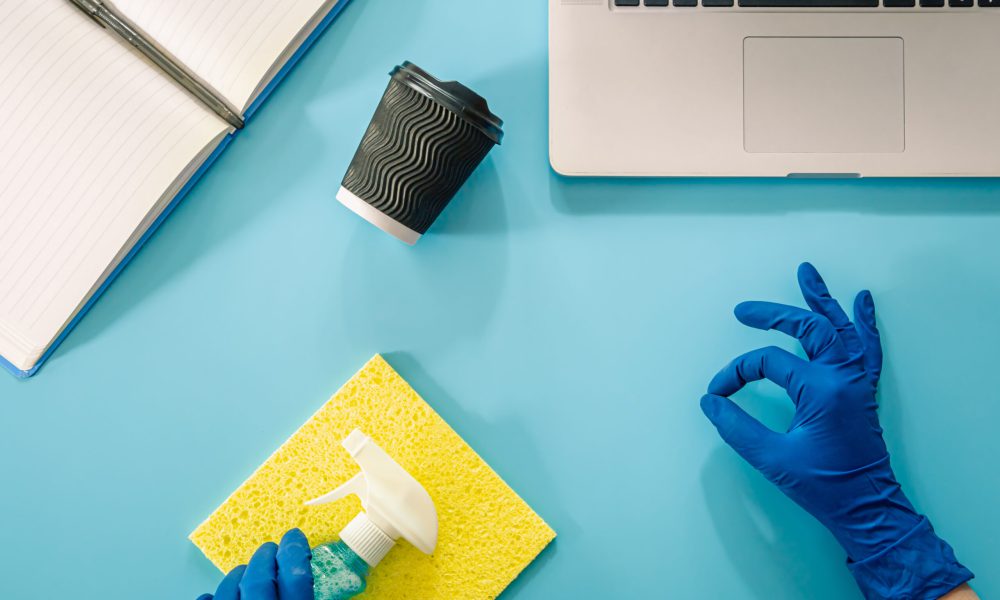 A hand in a rubber glove wipes a workplace with a laptop with a rag, flat lay.