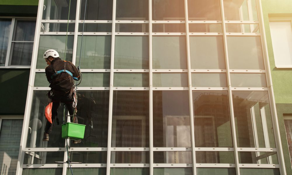 Concept of industry urban works. Industrial mountaineering worker in uniform hangs over residential facade building, washing exterior glazing. Rope access laborer hangs on wall of house. Copy space