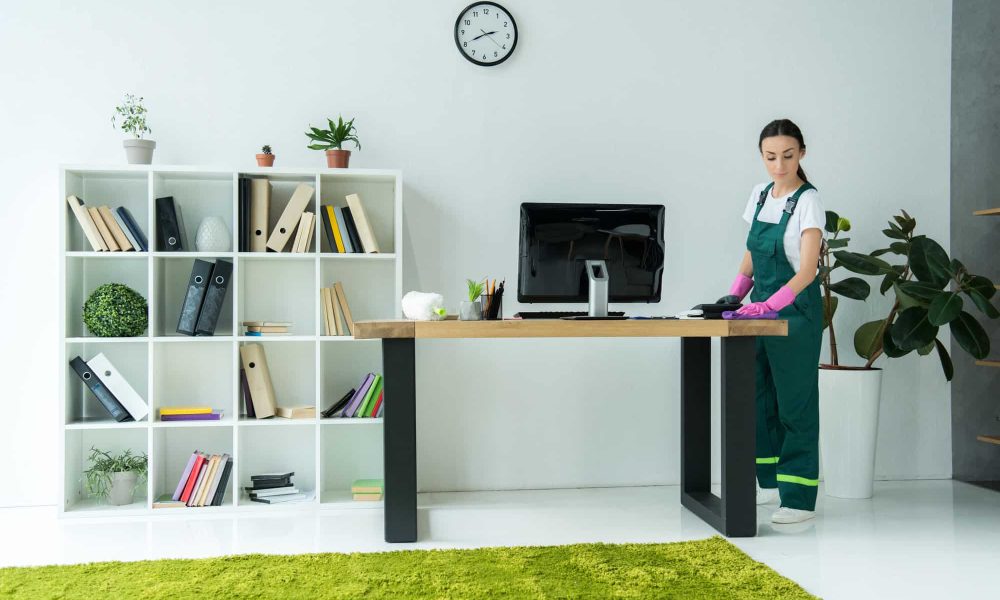 young-cleaning-company-worker-in-rubber-gloves-and-uniform-cleaning-desk-in-modern-office.jpg